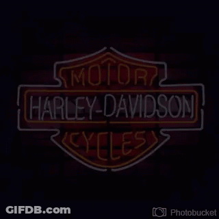 A Harley Davidson Neon Sign Is Lit Up On A Red Brick Wall . GIF