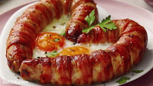 A Heart Shaped Sausage Wrapped In Bacon And Eggs On A White Plate . GIF