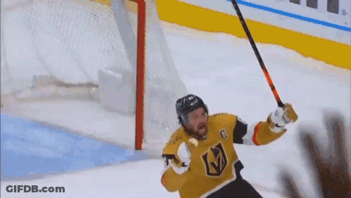 A Hockey Player Is Holding A Hockey Stick In The Air In Front Of A Goal . GIF