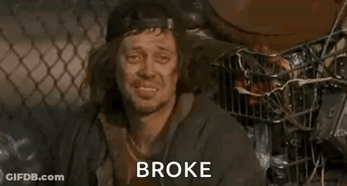 A Homeless Man Is Sitting In Front Of A Chain Link Fence With The Words `` Broke '' Written On His Face . GIF