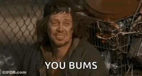 A Homeless Man Is Sitting In Front Of A Chain Link Fence With The Words `` You Bums '' Written On His Face . GIF