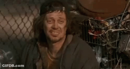 A Homeless Man Wearing A Baseball Cap And A Jacket Is Smiling While Sitting In Front Of A Chain Link Fence . GIF