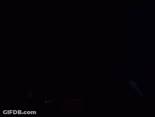 A Hooded Figure In A Dark Room With A Red And Black Mask GIF