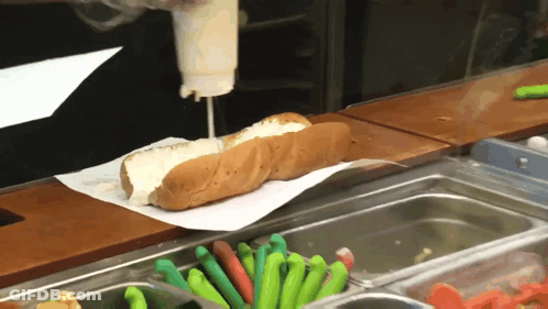 A Hot Dog With Mayonnaise Being Poured On It Sits On A Counter GIF