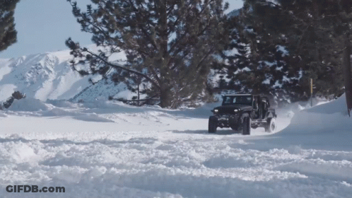 A Jeep Is Driving Through A Snowy Forest With Mountains In The Background GIF