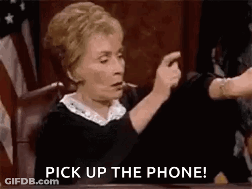A Judge Is Sitting At A Table In A Courtroom Holding A Cell Phone And Pointing At It . GIF
