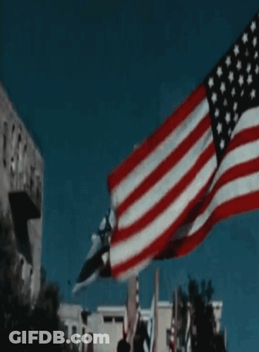A Large American Flag Is Flying In The Wind In Front Of A Building GIF