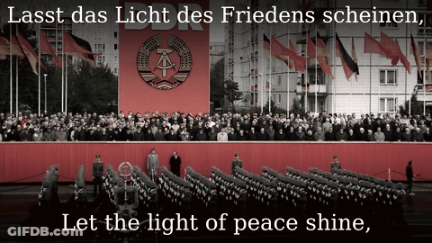 A Large Crowd Of People Are Gathered In Front Of A Large Red Banner That Says Ddr Let The Light Of Peace Shine GIF
