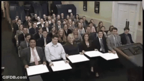 A Large Group Of People Are Posing For A Picture And One Of Them Is Holding A Piece Of Paper GIF