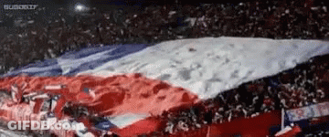 A Large Red , White And Blue Flag Is Being Waved By A Crowd Of People In A Stadium . GIF