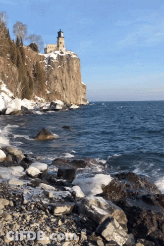 A Lighthouse Sits On Top Of A Rocky Cliff Overlooking The Ocean GIF