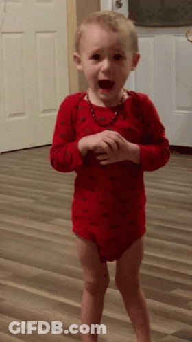 A Little Boy In A Red Bodysuit Is Standing On A Wood Floor GIF
