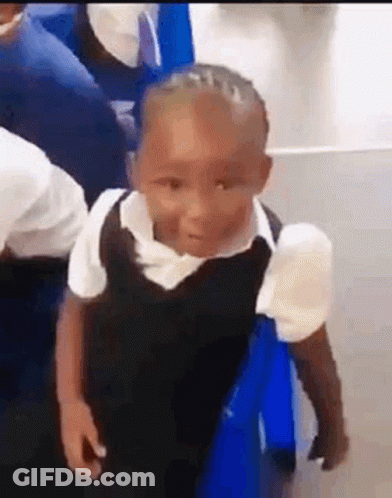 A Little Boy In A School Uniform Is Standing Next To A Blue Chair In A Classroom . GIF