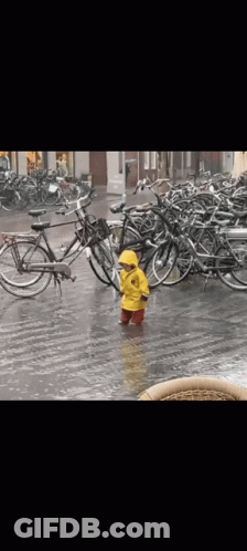 A Little Boy In A Yellow Raincoat Is Standing In The Rain In Front Of A Row Of Bikes . GIF