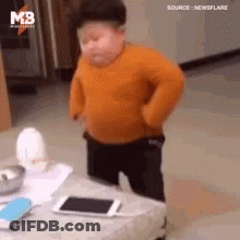 A Little Boy In An Orange Sweater Is Standing Next To A Table . GIF