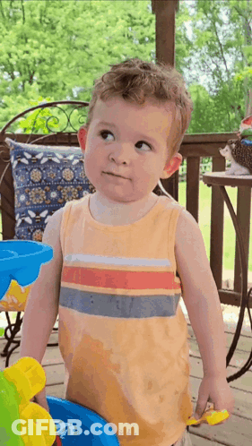A Little Boy In An Orange Tank Top Is Standing On A Porch Looking Up GIF