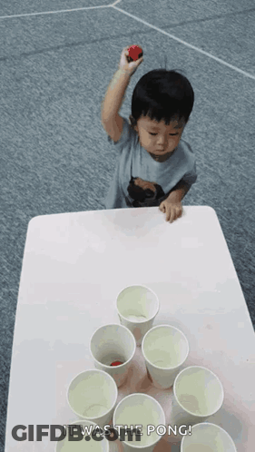 A Little Boy Is Playing Beer Pong With Cups And A Red Ball GIF
