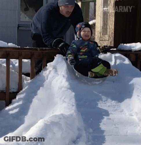 A Little Boy Is Sliding Down A Snowy Hill While A Man Stands Behind Him GIF