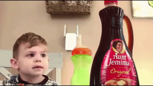 A Little Boy Is Standing In Front Of A Bottle Of Aunt Jemima Original Syrup GIF