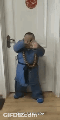 A Little Boy Is Standing In Front Of A Door Wearing A Blue Robe And A Necklace Of Beads . GIF