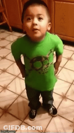 A Little Boy Is Standing On A Tiled Floor Wearing A Green Shirt . GIF