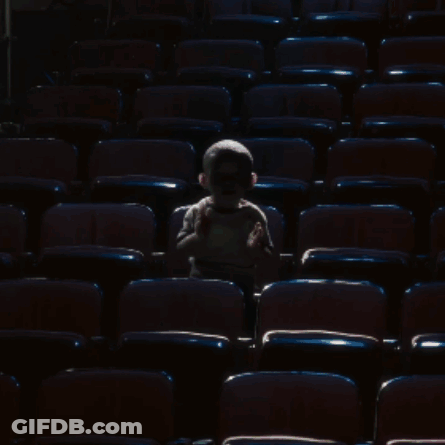 A Little Boy Sits In A Dark Auditorium With A Few Empty Seats GIF