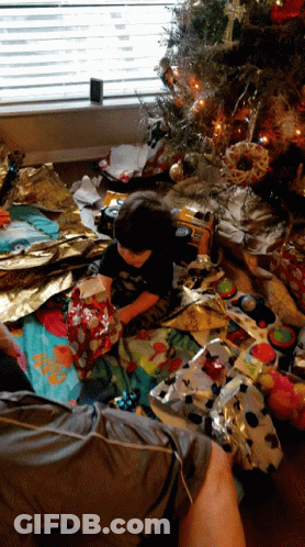 A Little Boy Sits On A Blanket In Front Of A Christmas Tree Opening A Present GIF