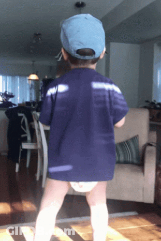 A Little Boy Wearing A Blue Hat And A Diaper Stands In A Living Room GIF