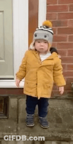 A Little Boy Wearing A Yellow Coat And A Hat Is Standing In Front Of A Brick Wall . GIF