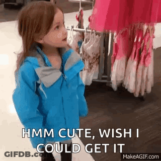 A Little Girl In A Blue Jacket Is Standing In Front Of A Rack Of Clothes And Talking To Someone . GIF