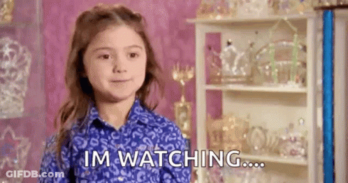 A Little Girl In A Blue Shirt Is Sitting In Front Of A Shelf With Trophies And Says `` Im Watching ... '' . GIF