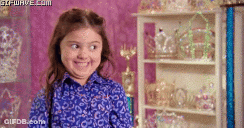 A Little Girl In A Blue Shirt Is Smiling In Front Of A Display Of Tiaras . GIF