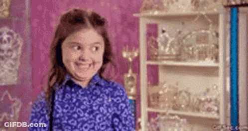 A Little Girl In A Blue Shirt Is Smiling In Front Of A Shelf Full Of Trophies . GIF