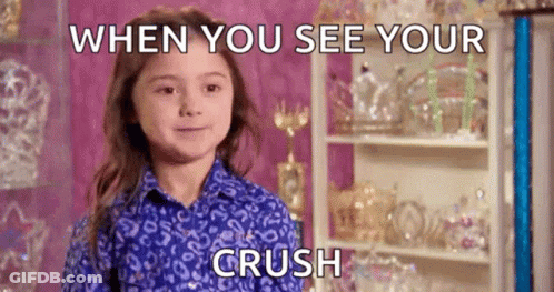 A Little Girl In A Blue Shirt Is Smiling While Standing In Front Of A Shelf With Trophies On It . GIF