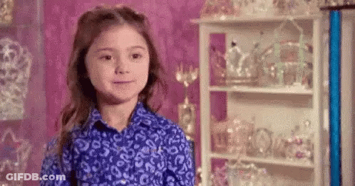 A Little Girl In A Blue Shirt Is Standing In Front Of A Shelf Filled With Crowns And Tiaras . GIF