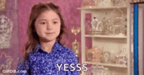 A Little Girl In A Blue Shirt Is Standing In Front Of A Shelf With A Shelf Full Of Trophies . GIF