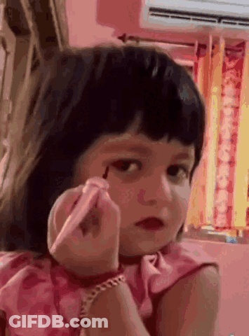A Little Girl In A Pink Dress Is Applying Makeup To Her Face GIF