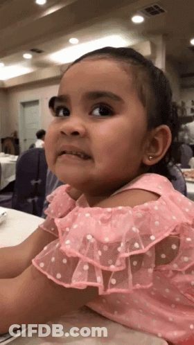 A Little Girl In A Pink Dress With White Polka Dots Is Sitting At A Table GIF
