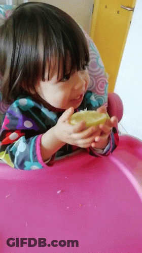 A Little Girl In A Pink High Chair Is Eating A Lemon GIF