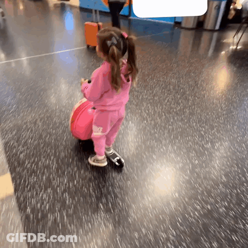 A Little Girl In A Pink Outfit Is Pulling A Suitcase GIF
