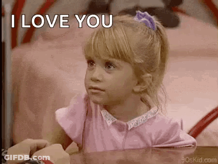 A Little Girl In A Pink Shirt Is Sitting At A Table With A Person And Says `` I Love You '' . GIF