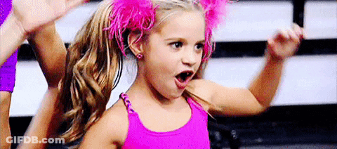 A Little Girl In A Pink Tank Top And Pink Feathers In Her Hair Is Making A Funny Face . GIF