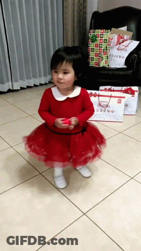 A Little Girl In A Red Dress Is Standing In Front Of A Chair And Christmas Presents GIF