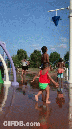 A Little Girl In A Red Top And Green Shorts Is Playing In A Water Fountain GIF