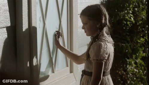 A Little Girl In A White Dress Is Looking At A Window GIF