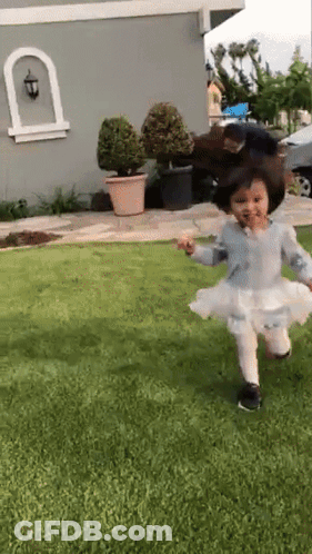 A Little Girl In A White Dress Is Running On The Grass In Front Of A House GIF