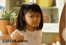 A Little Girl In A White Dress Is Sitting In Front Of A Window . GIF