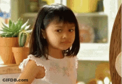 A Little Girl In A White Dress Is Standing In A Kitchen Looking At The Camera . GIF