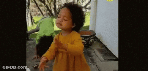 A Little Girl In A Yellow Dress Is Dancing In Front Of A Potted Plant And A Man In A Green Shirt . GIF