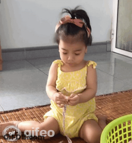 A Little Girl In A Yellow Dress Is Sitting On The Floor Playing With A Ribbon And A Basket With Gif Written On It GIF